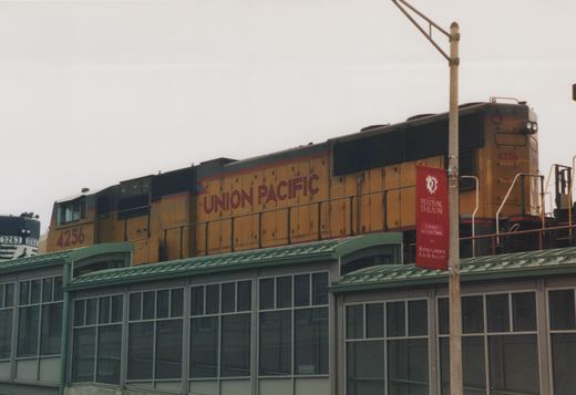 A yellow and black Union Pacific diesel locomotive numbered 4256 travels on an elevated track above a station platform, with a red banner for the Festival Theatre hanging on a nearby pole.