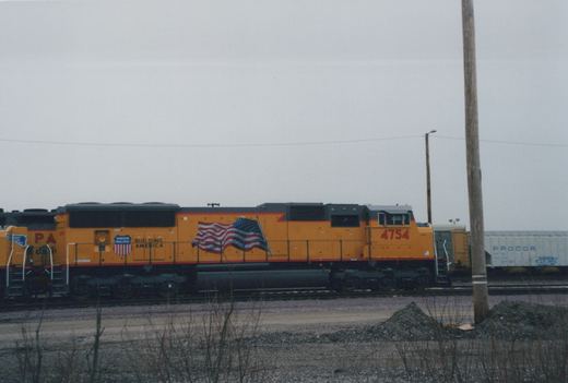A yellow Union Pacific locomotive numbered 4754 with an American flag graphic, standing on tracks in a yard with a white freight car in the background.