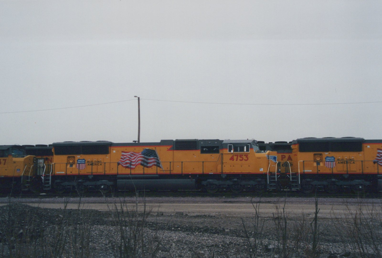 Side view of a yellow and black Union Pacific locomotive numbered 4753, with American flags painted on its side, standing in a gravel yard under a gray sky.