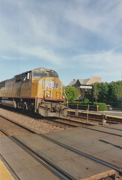 A yellow and gray Union Pacific locomotive numbered 4106 sits on the tracks at a station platform in Glen Ellyn under a blue sky with scattered clouds.