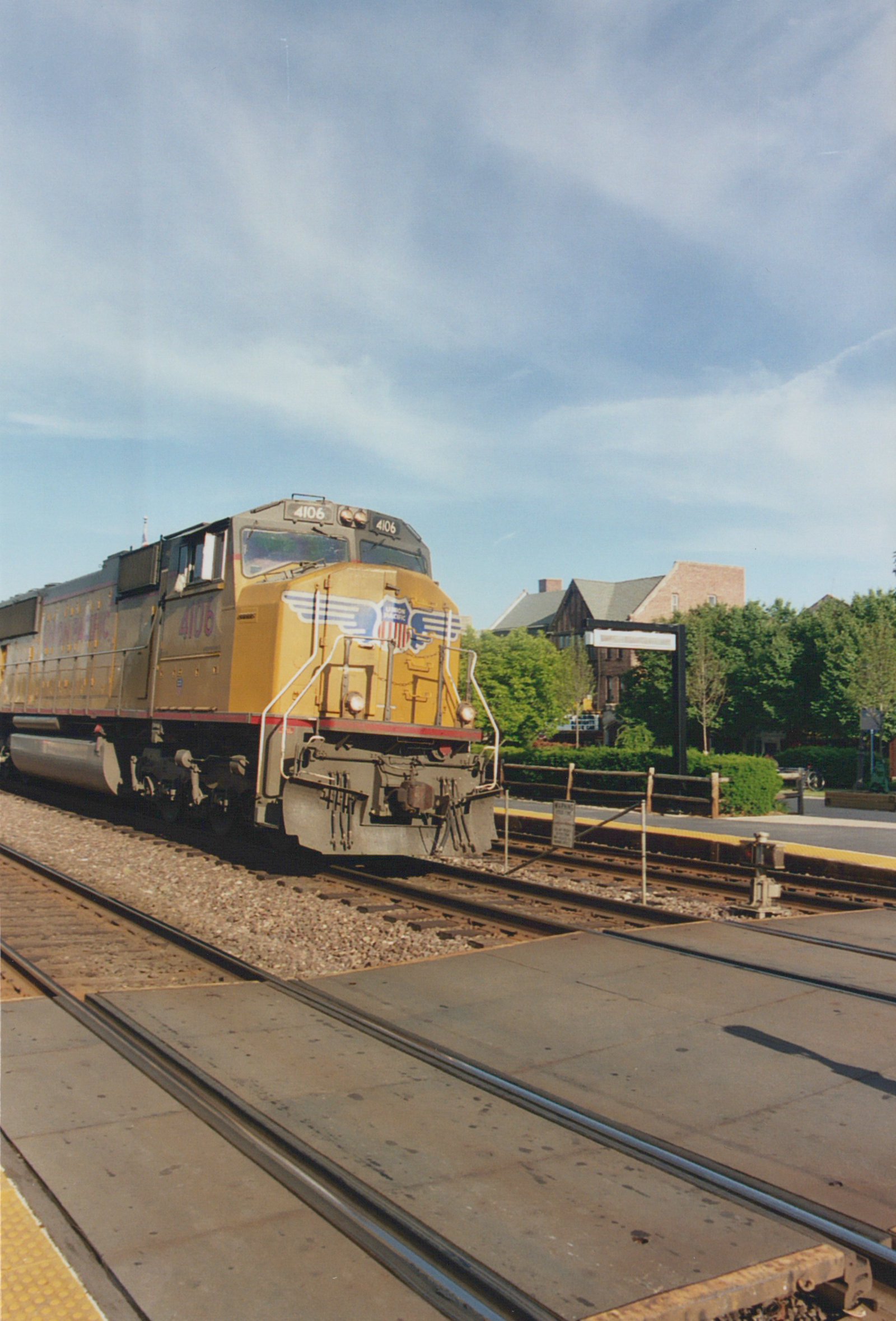 A yellow and gray Union Pacific locomotive numbered 4106 sits on the tracks at a station platform in Glen Ellyn under a blue sky with scattered clouds.