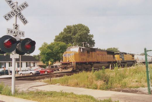 A yellow and black Union Pacific diesel locomotive with the number 4096 is moving on tracks at a grade crossing, with a railroad crossing sign in the foreground and several cars stopped on the road.