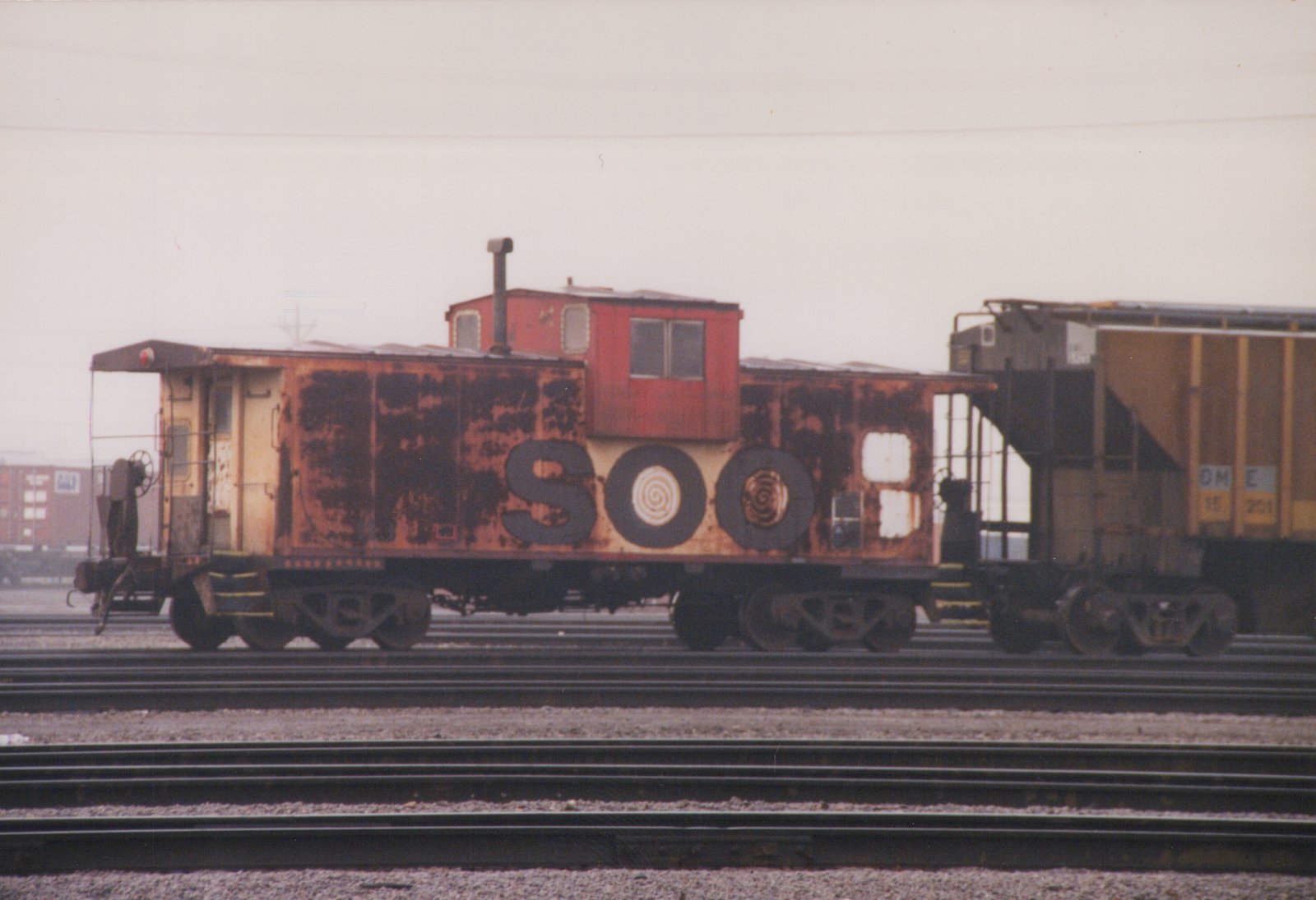 A rusty Soo Line caboose on multiple tracks in a rail yard under an overcast sky, with a freight car coupled behind it.