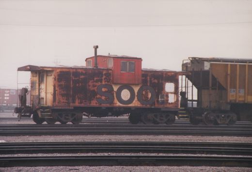 A rusty Soo Line caboose on multiple tracks in a rail yard under an overcast sky, with a freight car coupled behind it.
