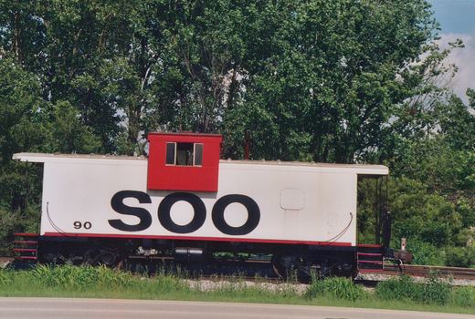 Side view of a white Soo Line caboose with red cupola, number 90, parked on railroad tracks next to a road with green trees in the background under a clear sky.