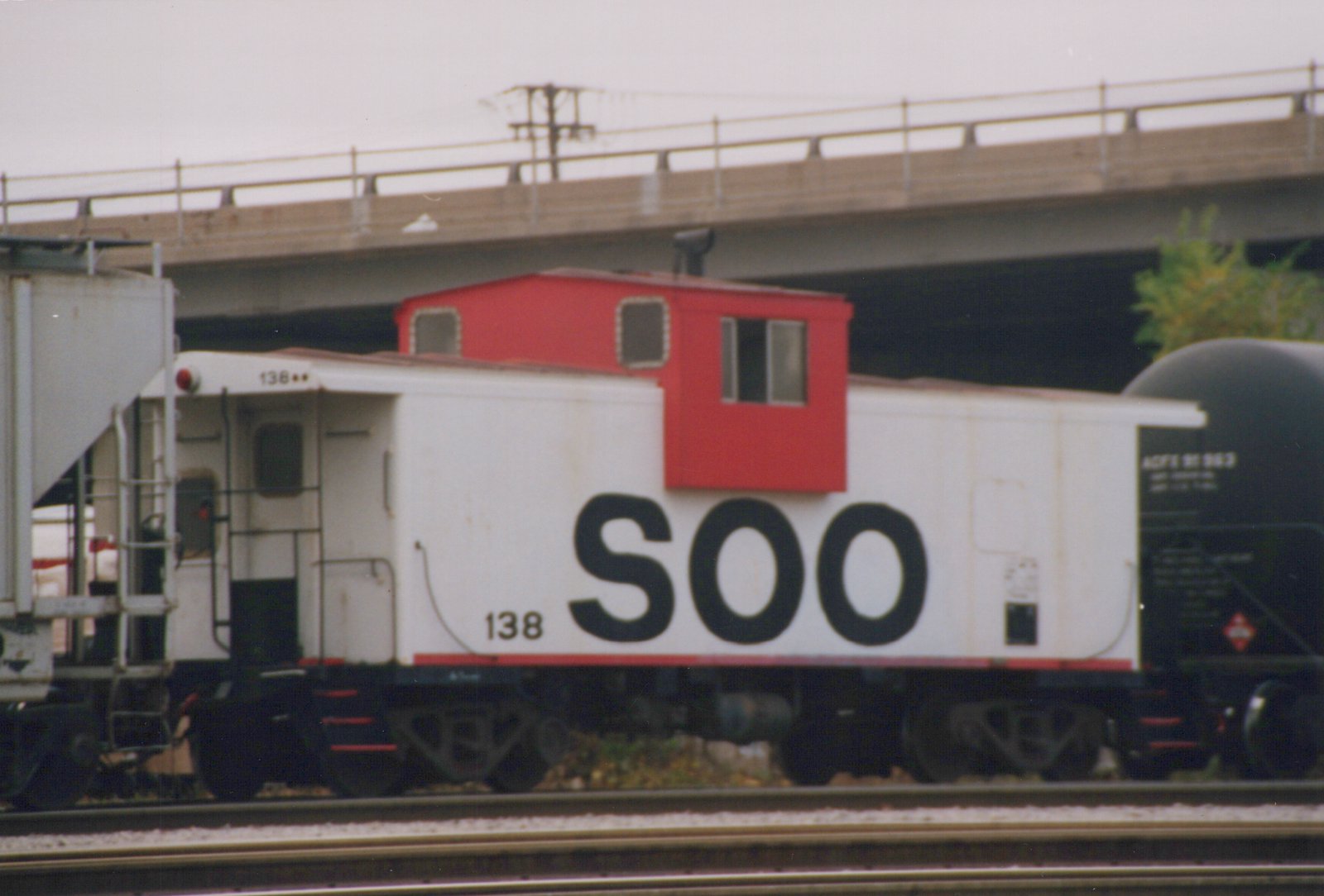 Side view of a white Soo Line caboose with number 138 and a red cupola, on train tracks under a concrete bridge with an ACFX tank car behind it.