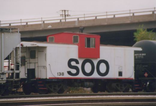 Side view of a white Soo Line caboose with number 138 and a red cupola, on train tracks under a concrete bridge with an ACFX tank car behind it.