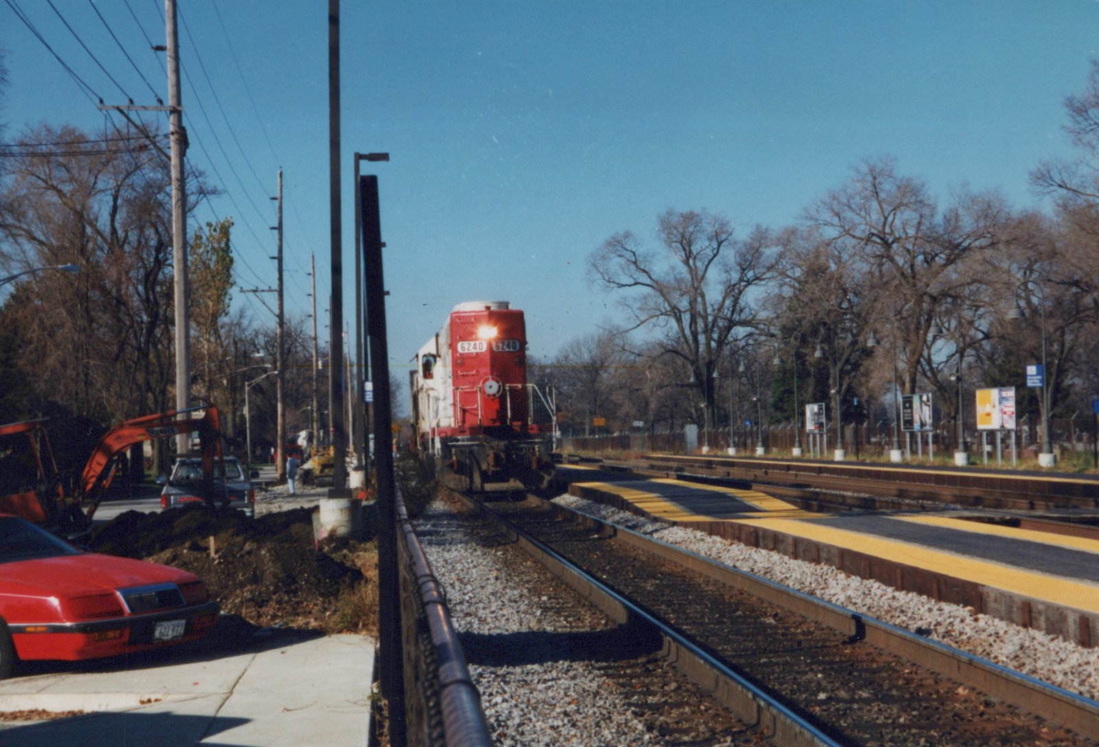 A red and white Soo Line locomotive numbered 6240 travels on tracks near a station platform with construction equipment and cars nearby on a sunny autumn day.
