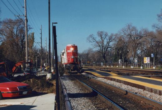 A red and white Soo Line locomotive numbered 6240 travels on tracks near a station platform with construction equipment and cars nearby on a sunny autumn day.