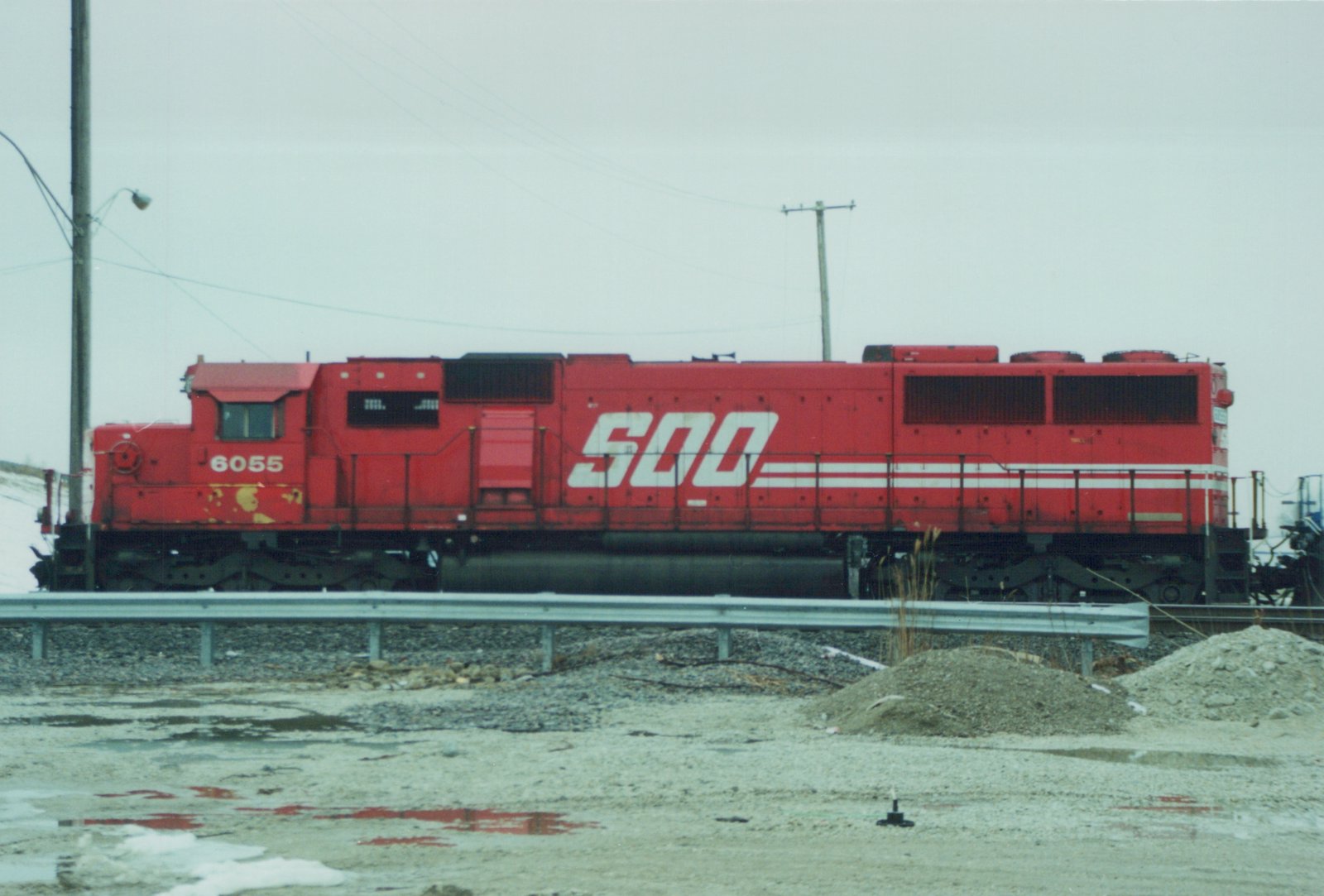 A bright red SOO Line diesel locomotive, number 6055, sits on the tracks beside a guardrail, with snow and puddles on the ground and a gray sky above.