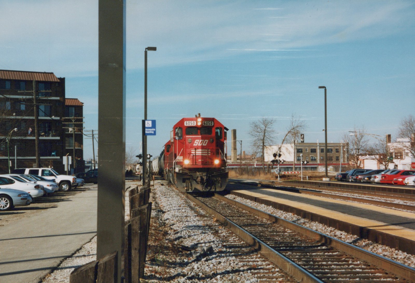 A red Soo Line locomotive number 6050 is moving on a set of tracks toward a station platform, with parked cars and buildings in the background on a clear winter day.