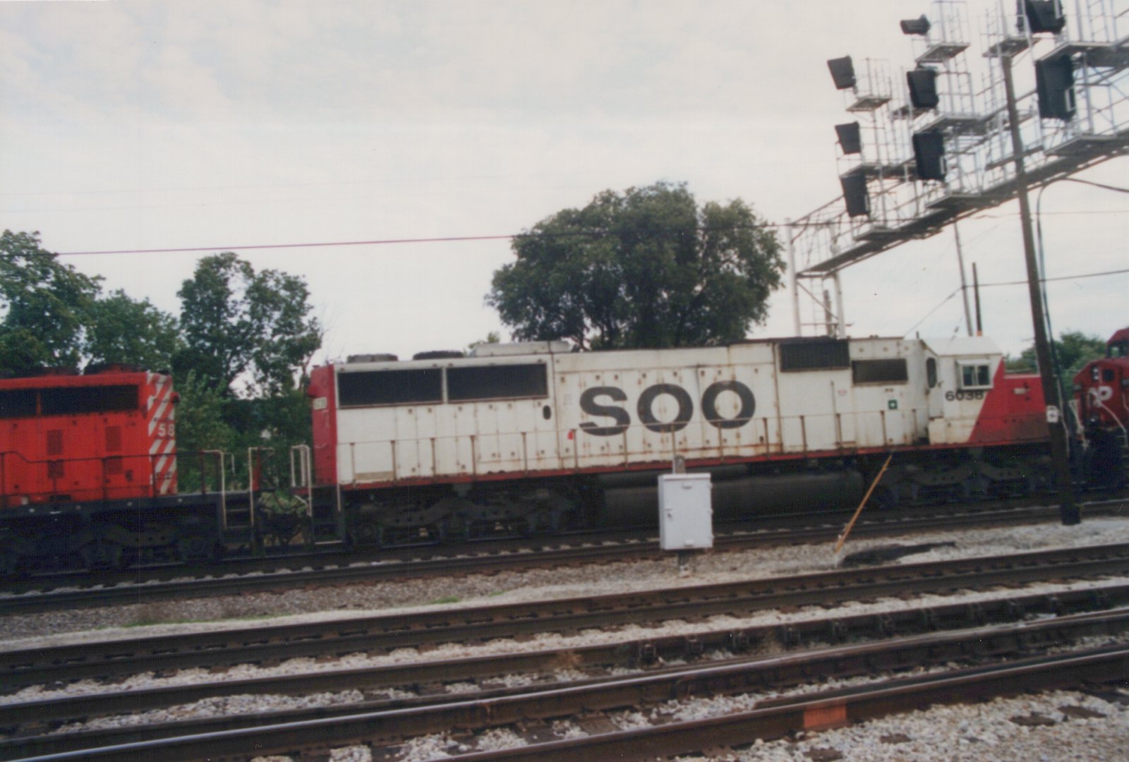 A white and red SOO Line diesel locomotive numbered 6038 is parked on tracks in a rail yard, with another red locomotive partially visible beside it.