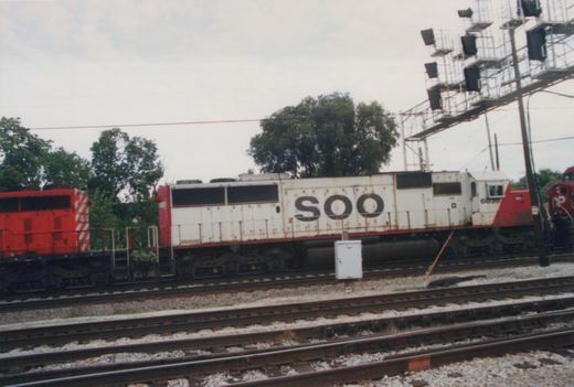 A white and red SOO Line diesel locomotive numbered 6038 is parked on tracks in a rail yard, with another red locomotive partially visible beside it.
