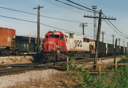 A red and white SOO Line locomotive numbered 6020 pulls a freight train of boxcars and hoppers along a railroad track, with telephone poles and green bushes in the foreground.