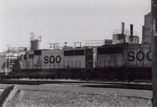 A black and white photo of a Soo Line locomotive numbered 6005, a large diesel engine, parked beside a freight car on railroad tracks in a yard with a cloudy sky.