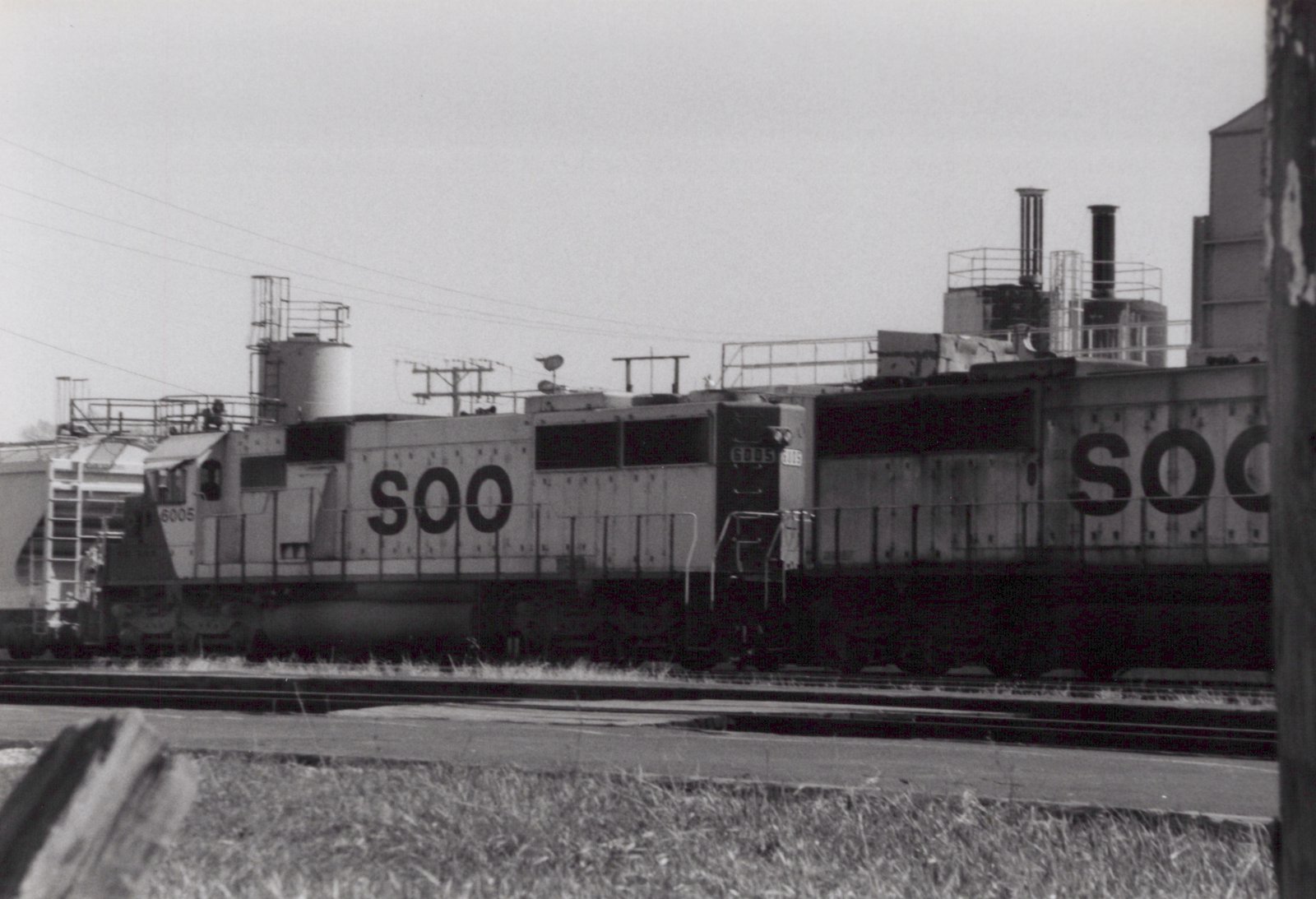 A black and white photo of a Soo Line locomotive numbered 6005, a large diesel engine, parked beside a freight car on railroad tracks in a yard with a cloudy sky.