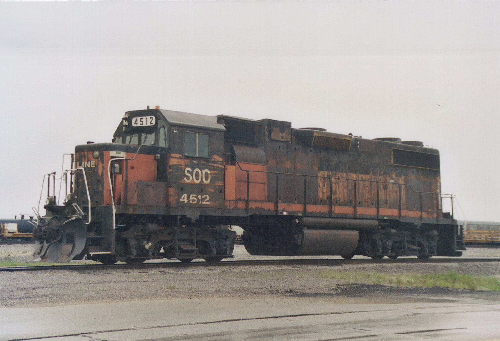 A side view of a weathered black and orange SOO Line diesel locomotive number 4512, sitting on tracks in a yard with other freight cars visible in the background under an overcast sky.