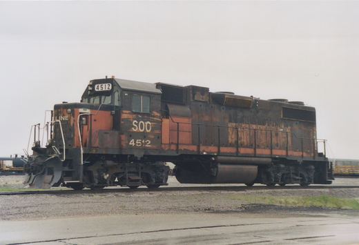 A side view of a weathered black and orange SOO Line diesel locomotive number 4512, sitting on tracks in a yard with other freight cars visible in the background under an overcast sky.