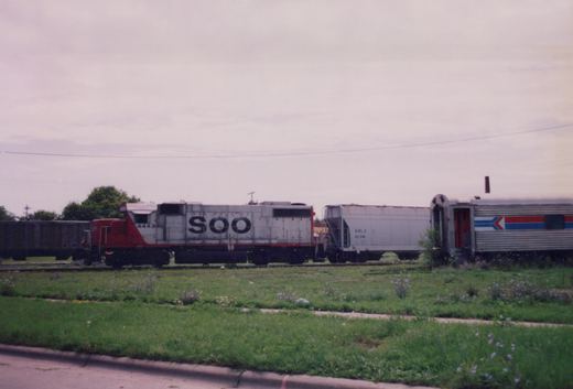 A side view of a Soo Line diesel locomotive with the number 4443, pulling a freight car and an Amtrak passenger car, parked on tracks next to a grassy roadside on an overcast day.