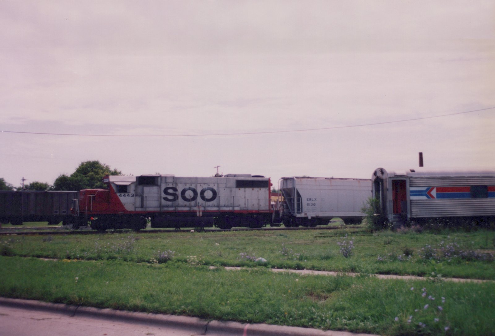 A side view of a Soo Line diesel locomotive with the number 4443, pulling a freight car and an Amtrak passenger car, parked on tracks next to a grassy roadside on an overcast day.
