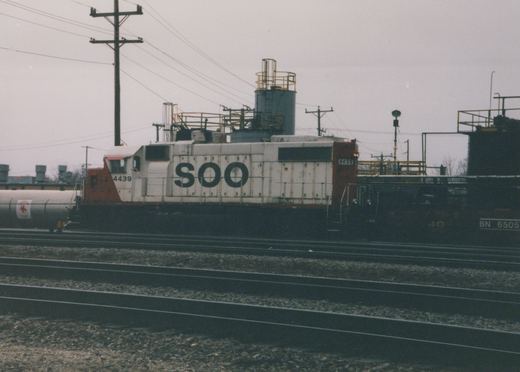 Side view of a white and orange SOO Line diesel locomotive numbered 4439 on multiple tracks in a rail yard, with a black Burlington Northern freight car nearby under an overcast sky.