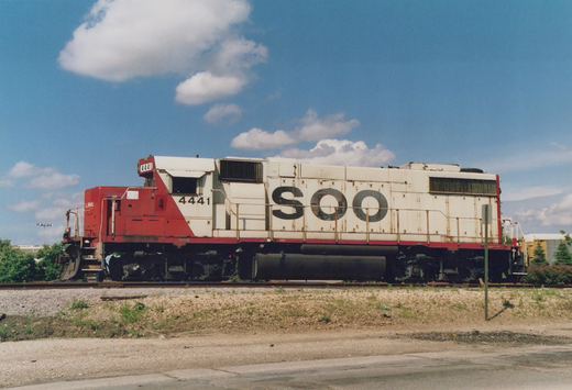 A side view of a red, white, and black Soo Line diesel locomotive, number 4441, standing on railway tracks under a blue sky with white clouds.