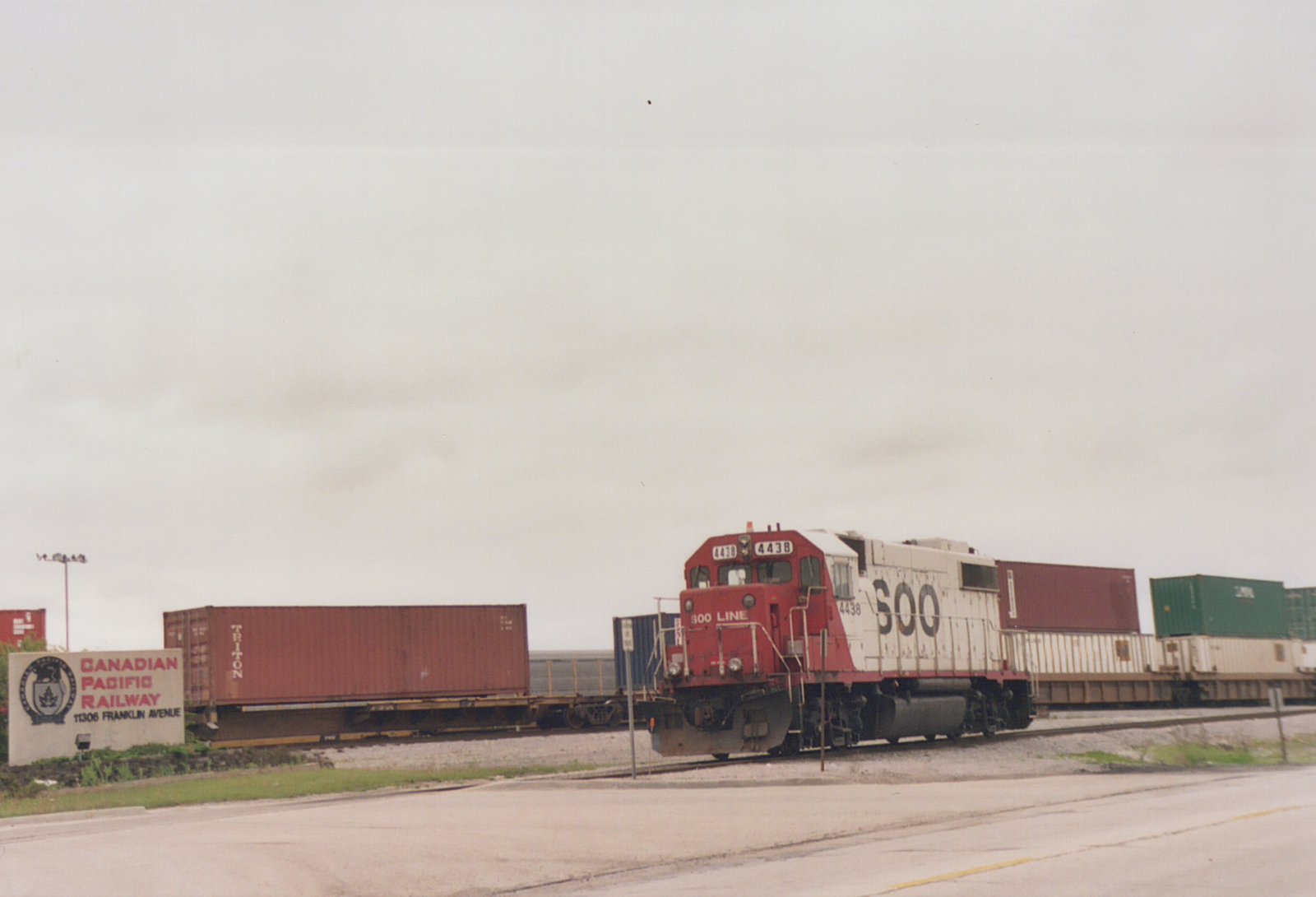 A red and white SOO Line locomotive with the number 4438 on its side pulls a line of freight containers on a railroad track next to a road, with a Canadian Pacific Railway sign visible in the background under an overcast sky.
