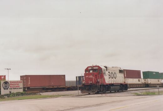 A red and white SOO Line locomotive with the number 4438 on its side pulls a line of freight containers on a railroad track next to a road, with a Canadian Pacific Railway sign visible in the background under an overcast sky.