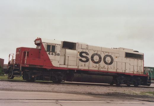 A side view of a white and red SOO Line diesel locomotive numbered 4438, parked on rail tracks in a gravel yard with a green boxcar partially visible in the background.
