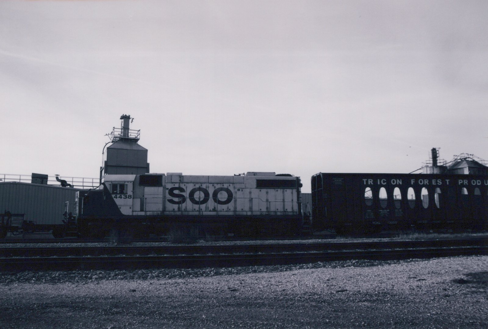 A black and white photograph of a SOO Line locomotive numbered 4438, parked beside a dark freight car with partial lettering, in a gravel yard with industrial buildings in the background.