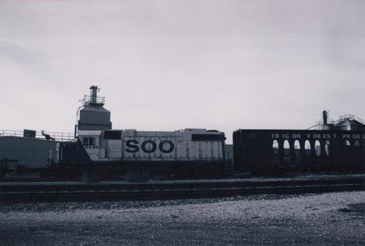 A black and white photograph of a SOO Line locomotive numbered 4438, parked beside a dark freight car with partial lettering, in a gravel yard with industrial buildings in the background.