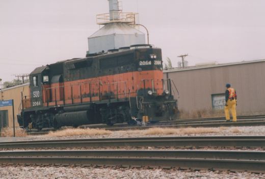 A black and orange SOO Line diesel locomotive number 2064 sits on railroad tracks with a person in safety gear nearby, with a sign reading Mannheim in the background.