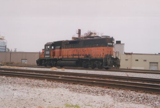 A rusted orange and black Soo Line GP38 locomotive, number 2064, sits on a set of tracks in an industrial area under an overcast sky.