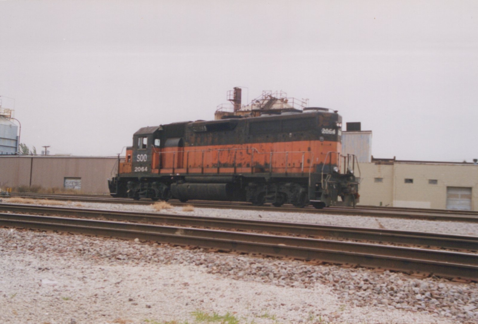 A rusted orange and black Soo Line GP38 locomotive, number 2064, sits on a set of tracks in an industrial area under an overcast sky.