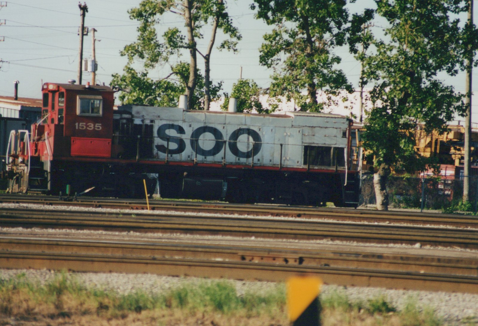 A red and silver Soo Line diesel locomotive, number 1535, stands on tracks in a yard with trees and other rail equipment in the background.