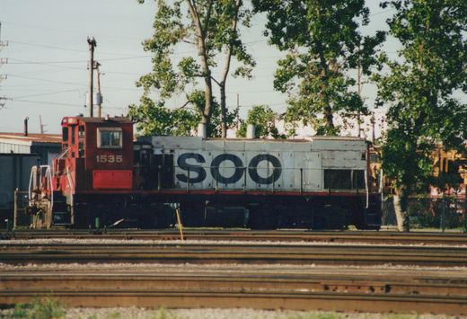 A red and white SOO Line diesel locomotive numbered 1535 sits on tracks in a rail yard with trees and overhead wires in the background.