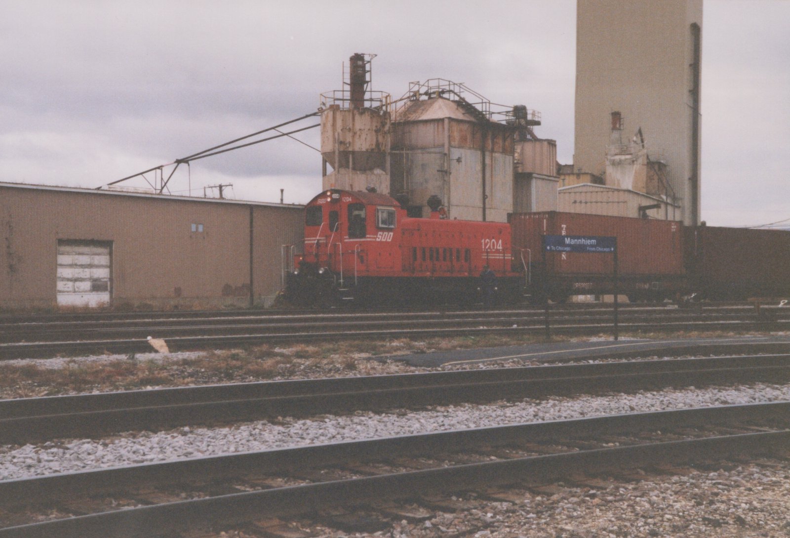 A red diesel locomotive labeled '1204' and '500' is coupled to a brown container car on multiple tracks in an industrial rail yard, with a station sign reading 'Mannhiem' visible.