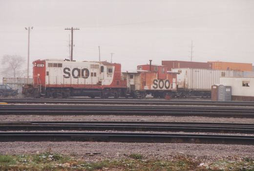 Side view of a red and white SOO diesel locomotive numbered 414, coupled to a matching SOO caboose numbered 102, in a multi-track railroad yard with shipping containers in the background on a cloudy day.