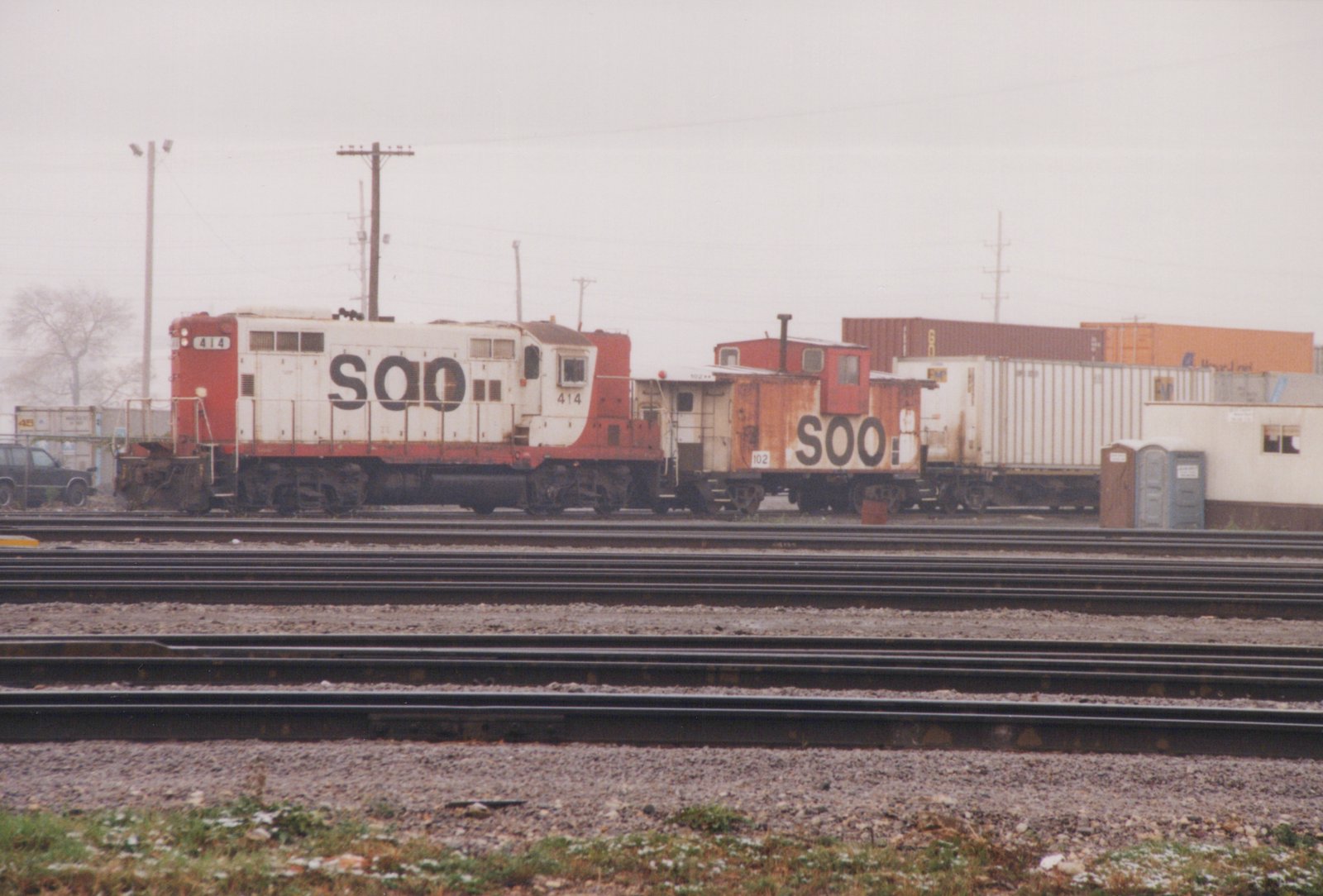 Side view of a red and white SOO diesel locomotive numbered 414, coupled to a matching SOO caboose numbered 102, in a multi-track railroad yard with shipping containers in the background on a cloudy day.