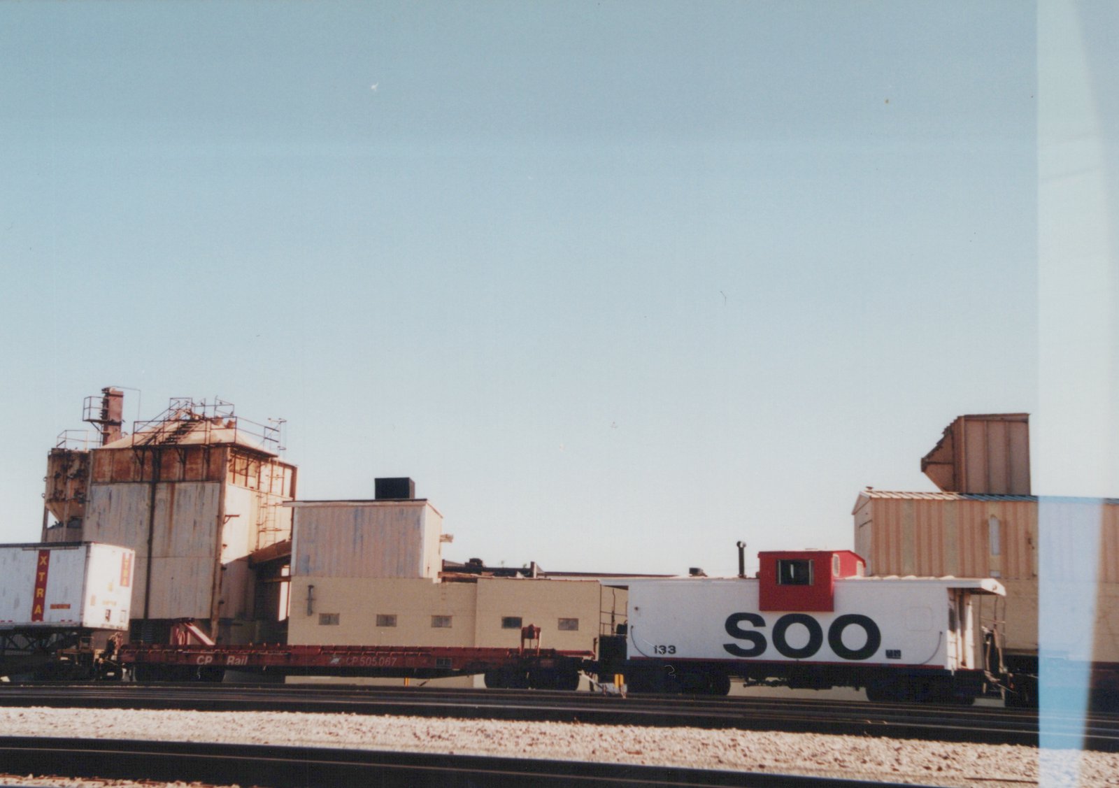 A white Soo Line caboose with the number 133 is parked on railroad tracks next to a red Canadian Pacific flatcar with the number CP 505067, which is carrying a white XTRA trailer, with industrial buildings in the background.