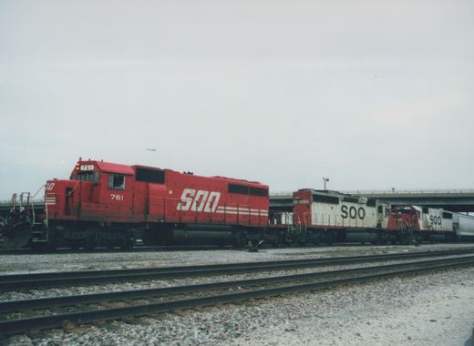 A side view of three Soo Line freight locomotives, one red SD40-2 and two white GP38s, on tracks in a yard with an overpass behind them under a gray sky.