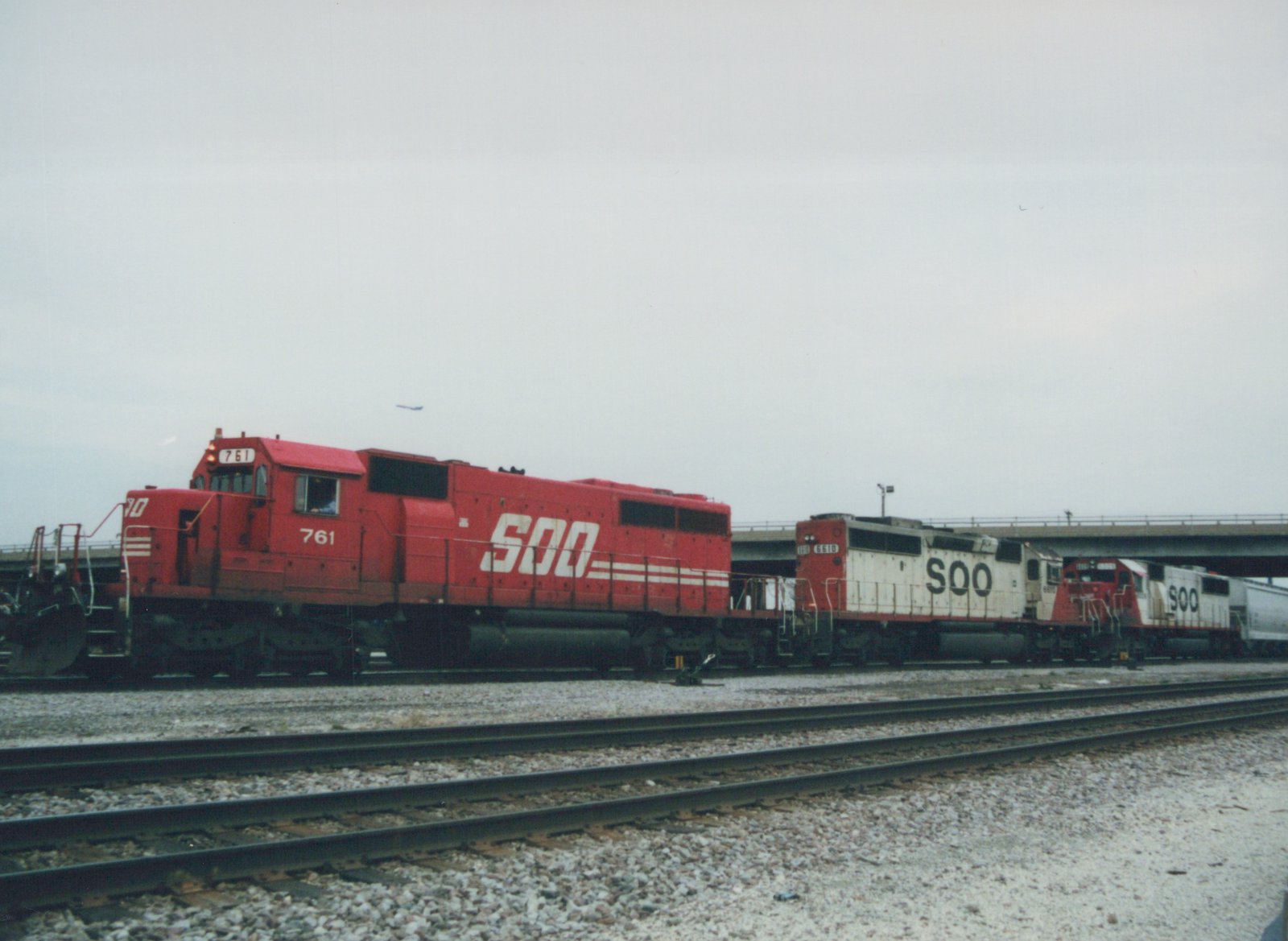 A side view of three Soo Line freight locomotives, one red SD40-2 and two white GP38s, on tracks in a yard with an overpass behind them under a gray sky.
