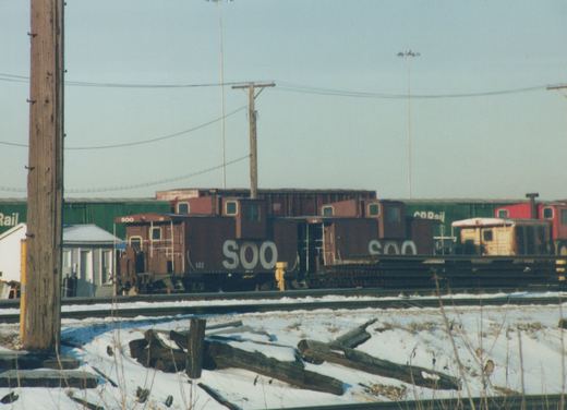 A maroon SOO Line caboose numbered 141 is parked on tracks in a snow-covered yard, with other freight cars in the background.