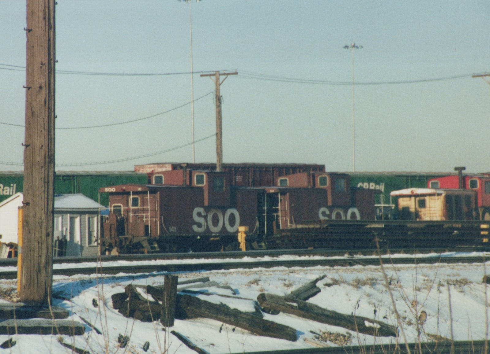 A maroon SOO Line caboose numbered 141 is parked on tracks in a snow-covered yard, with other freight cars in the background.
