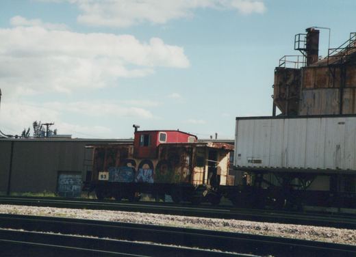 A side view of a rusty, graffiti-covered red caboose numbered 93 on railroad tracks, with a white semi-trailer on a flatcar beside it under a partly cloudy sky.