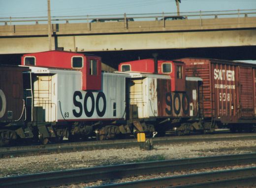A side view of two SOO Line cabooses, 63 and 94, coupled to a Southern Railway boxcar, on tracks under a concrete overpass on a clear day.