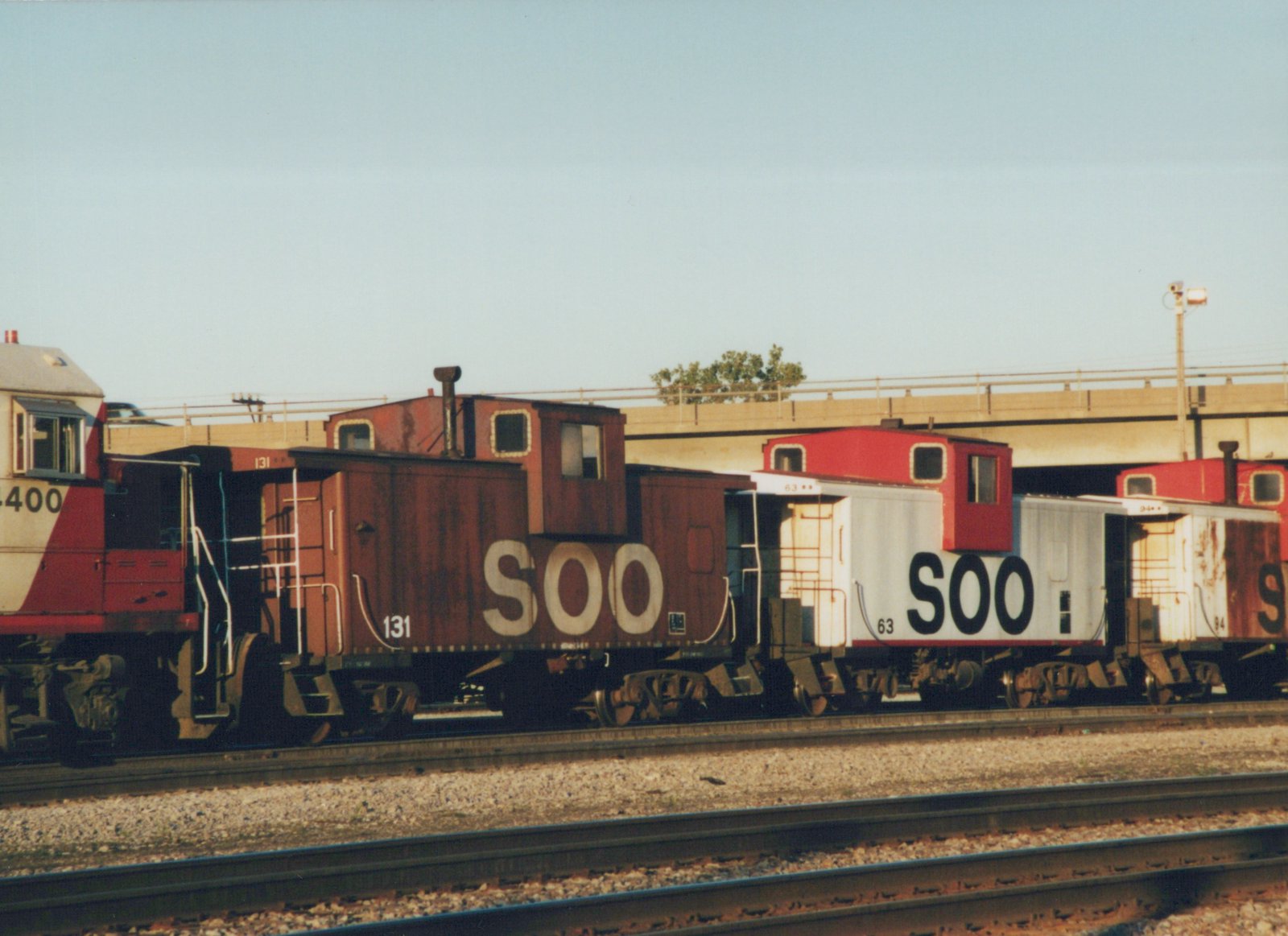 A side view of a Soo Line train with a locomotive and three cabooses on a clear day, with a bridge in the background.