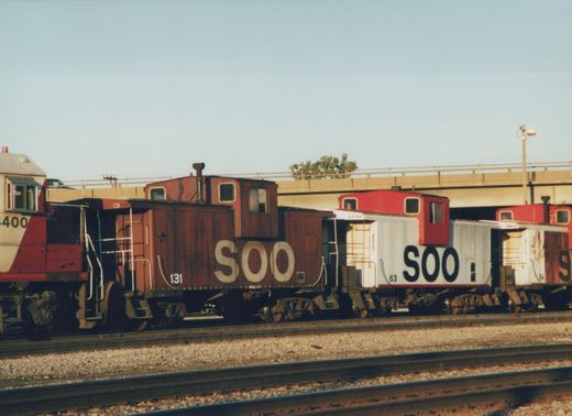 A side view of a Soo Line train with a locomotive and three cabooses on a clear day, with a bridge in the background.
