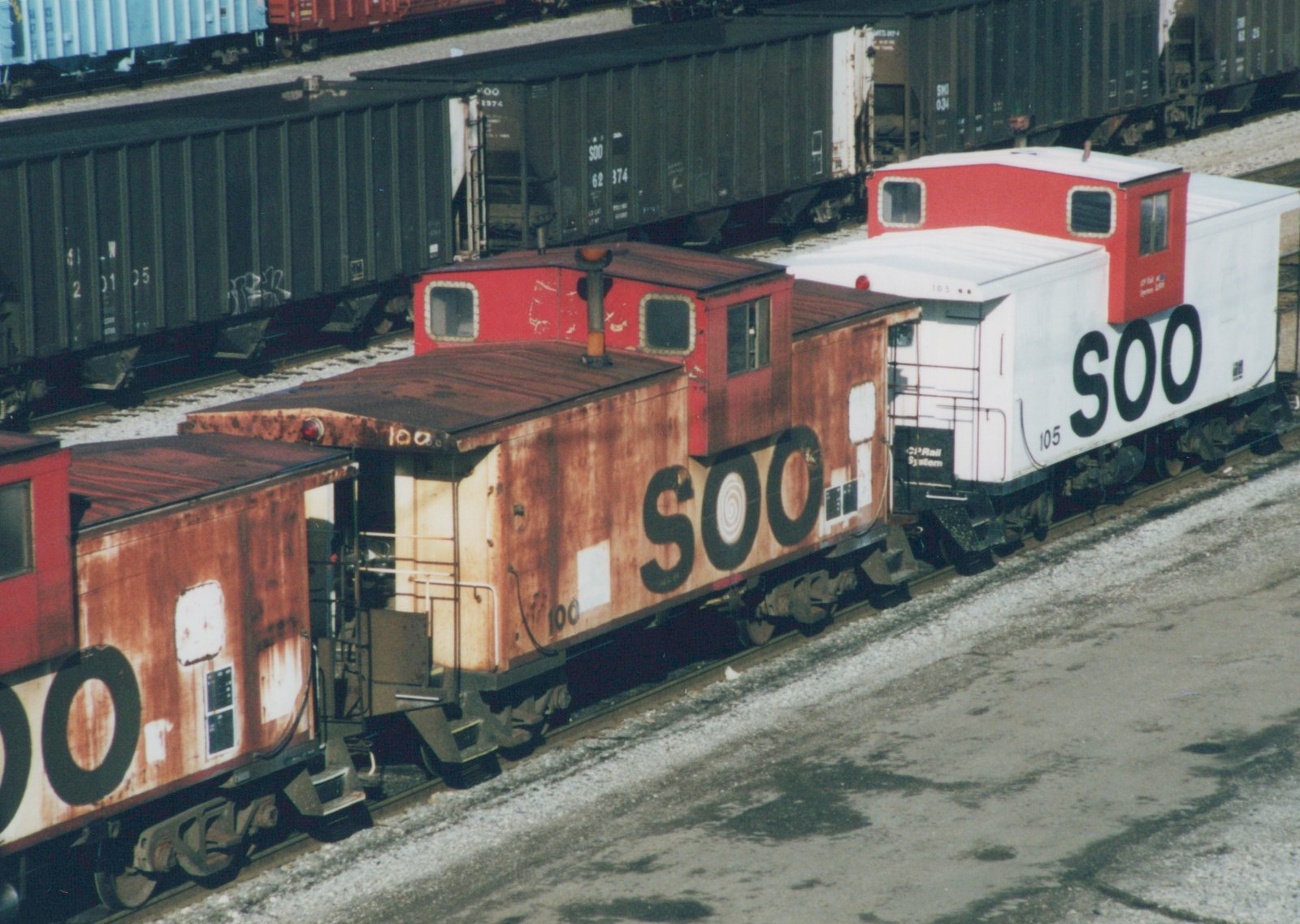 Two cabooses, one rusty brown and one white with red cupolas, both lettered SOO, are coupled together in a railyard with snow on the ground and other freight cars behind them.