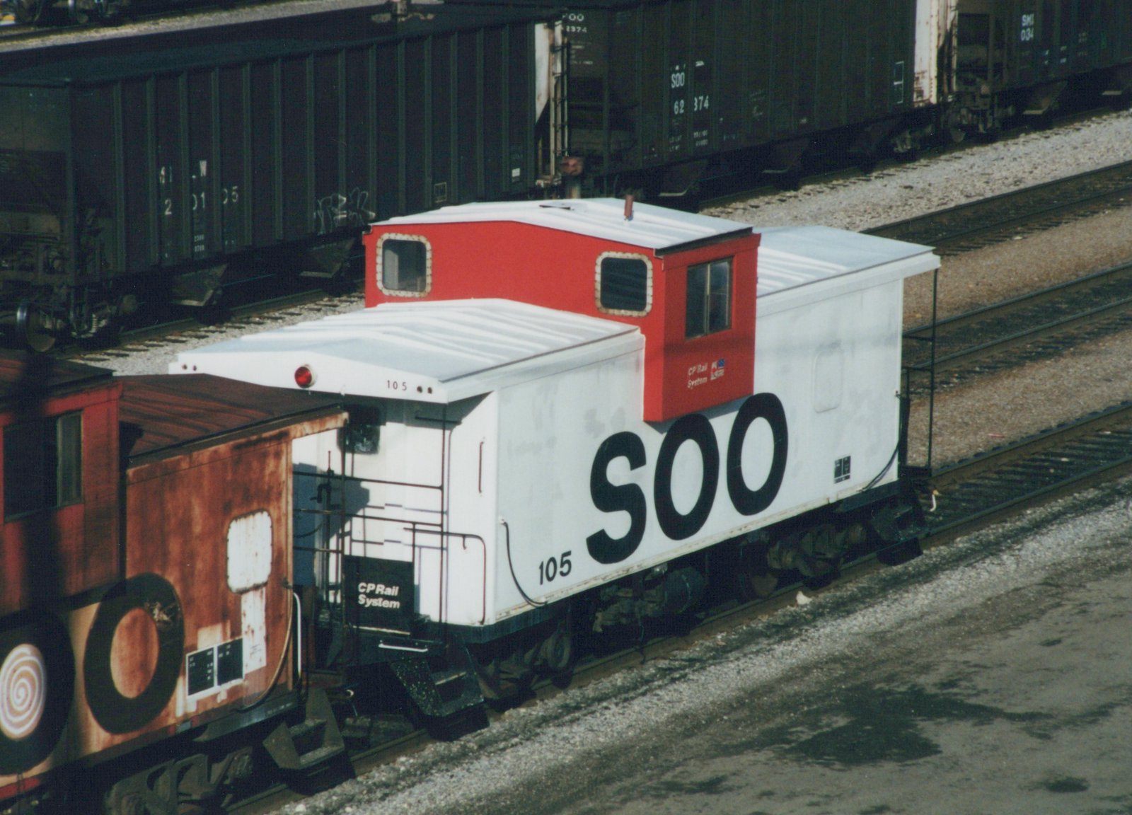 A white caboose with large black 'SOO' lettering and a red cupola, numbered 105, sitting on railroad tracks in a yard with dark green freight cars behind it.